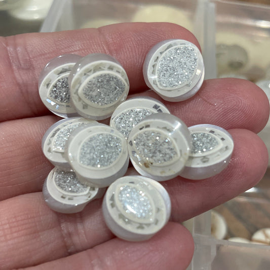 Hand holding small round decorative items with glittery surface, blurred container of similar items in the background.
