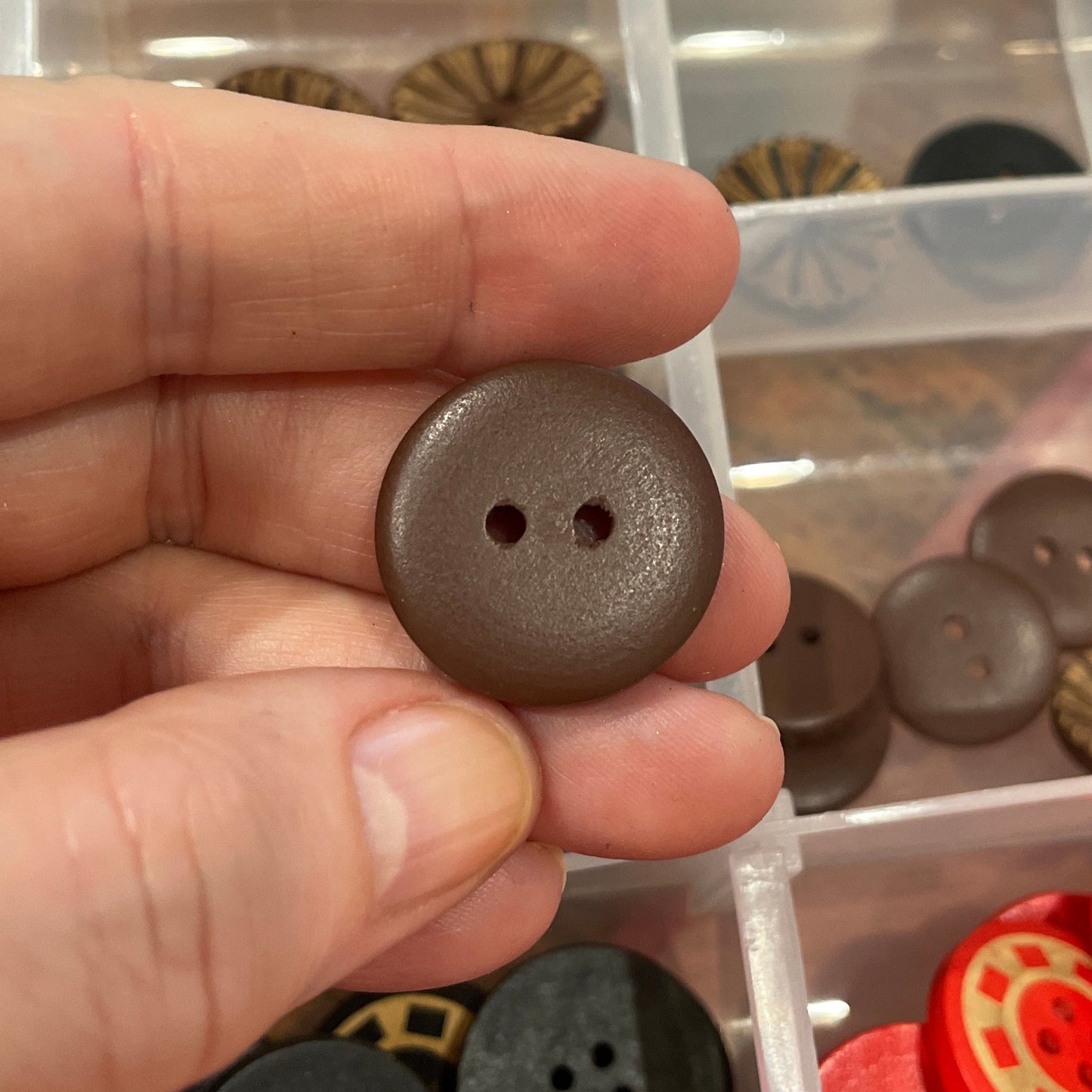 Hand holding a brown button with a clear container of assorted buttons in the background