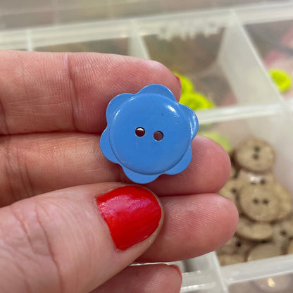 Hand holding a blue button with a flower design in front of a container of buttons.