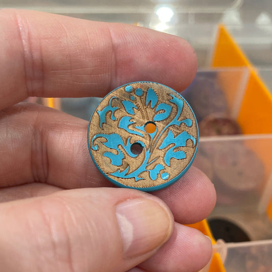 Hand holding a decorative button with intricate design, against a background of a button organizer.