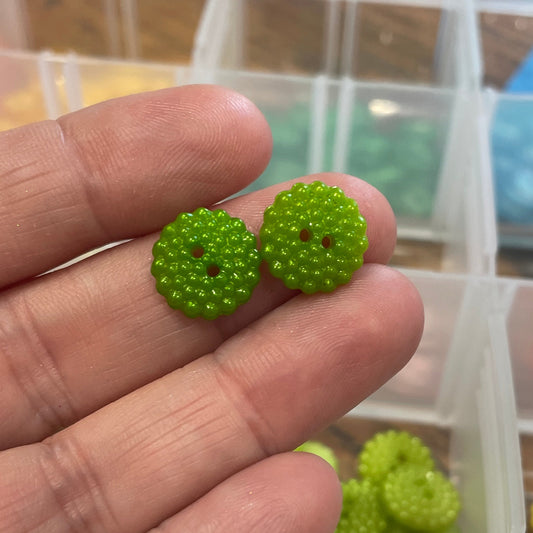 Green button earrings held between fingers with a container of buttons in the background