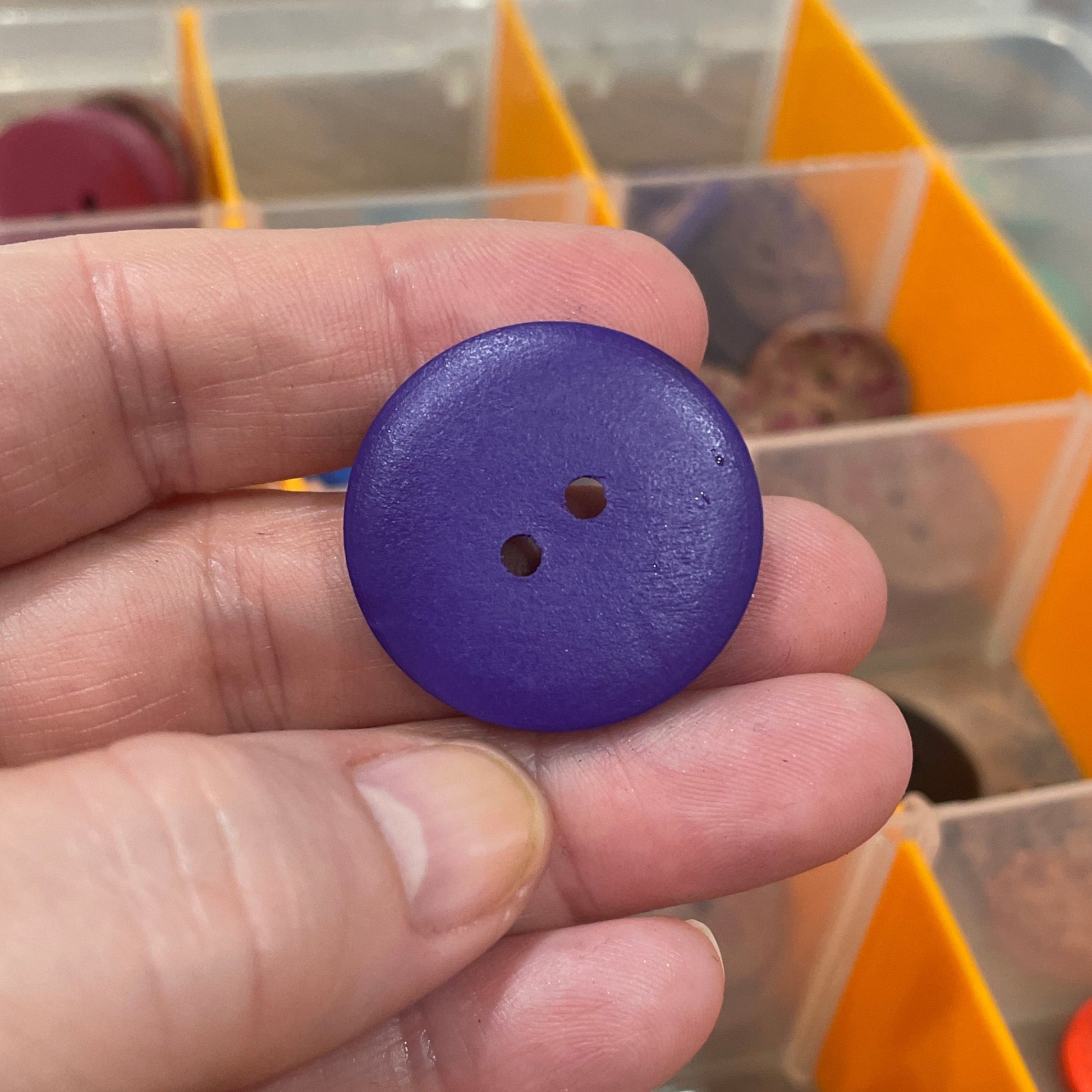 Hand holding a purple button with a button collection container in the background
