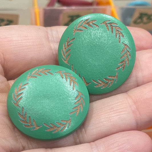 Two green buttons with gold patterns held in front of a container of assorted buttons.