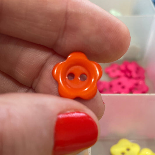 Hand holding an orange flower-shaped button with a container of colorful buttons in the background.