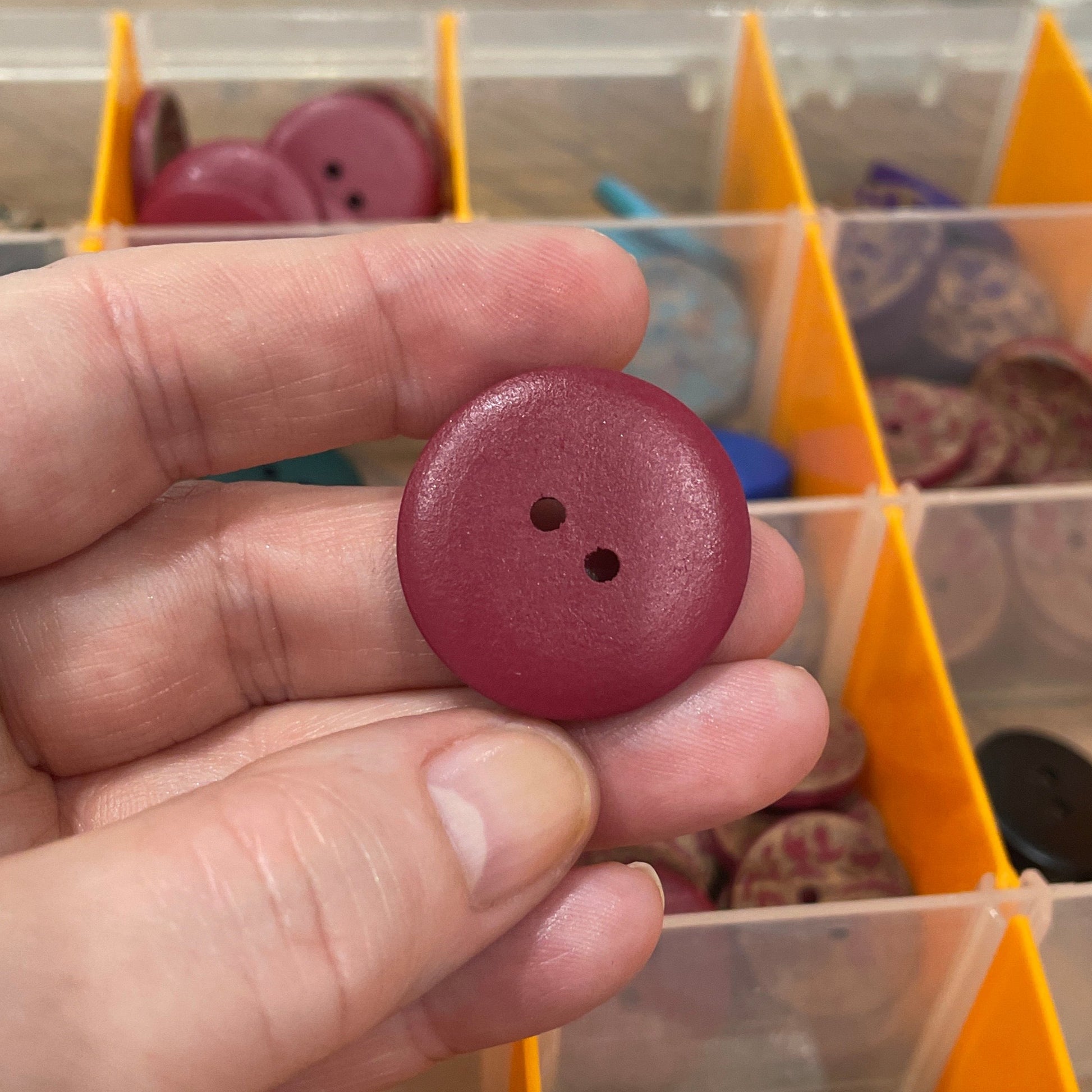 Hand holding a maroon button with a background of button storage containers.