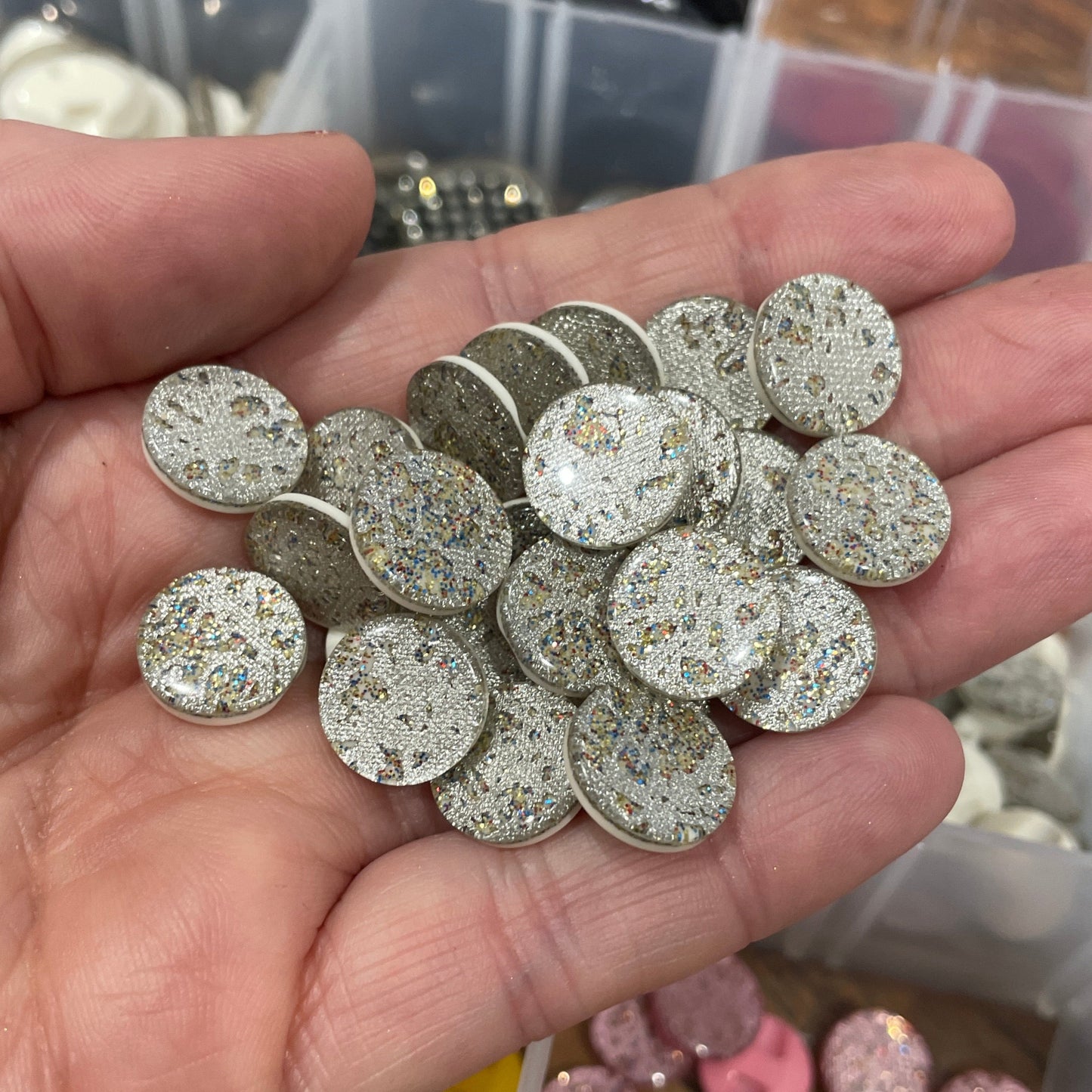 Hand holding silver embellishments with a container of colorful embellishments in the background