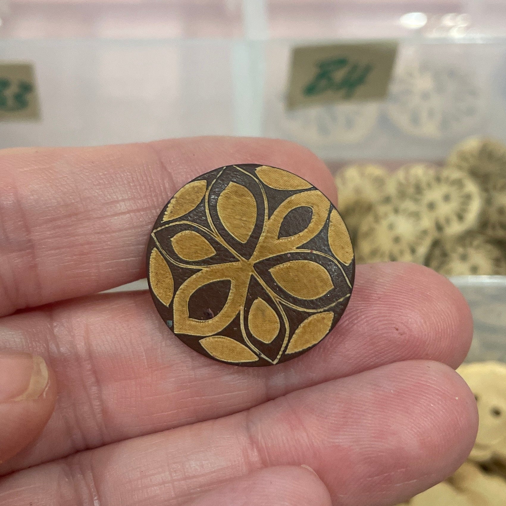 Hand holding a decorative button with a floral pattern in front of a container of assorted buttons.