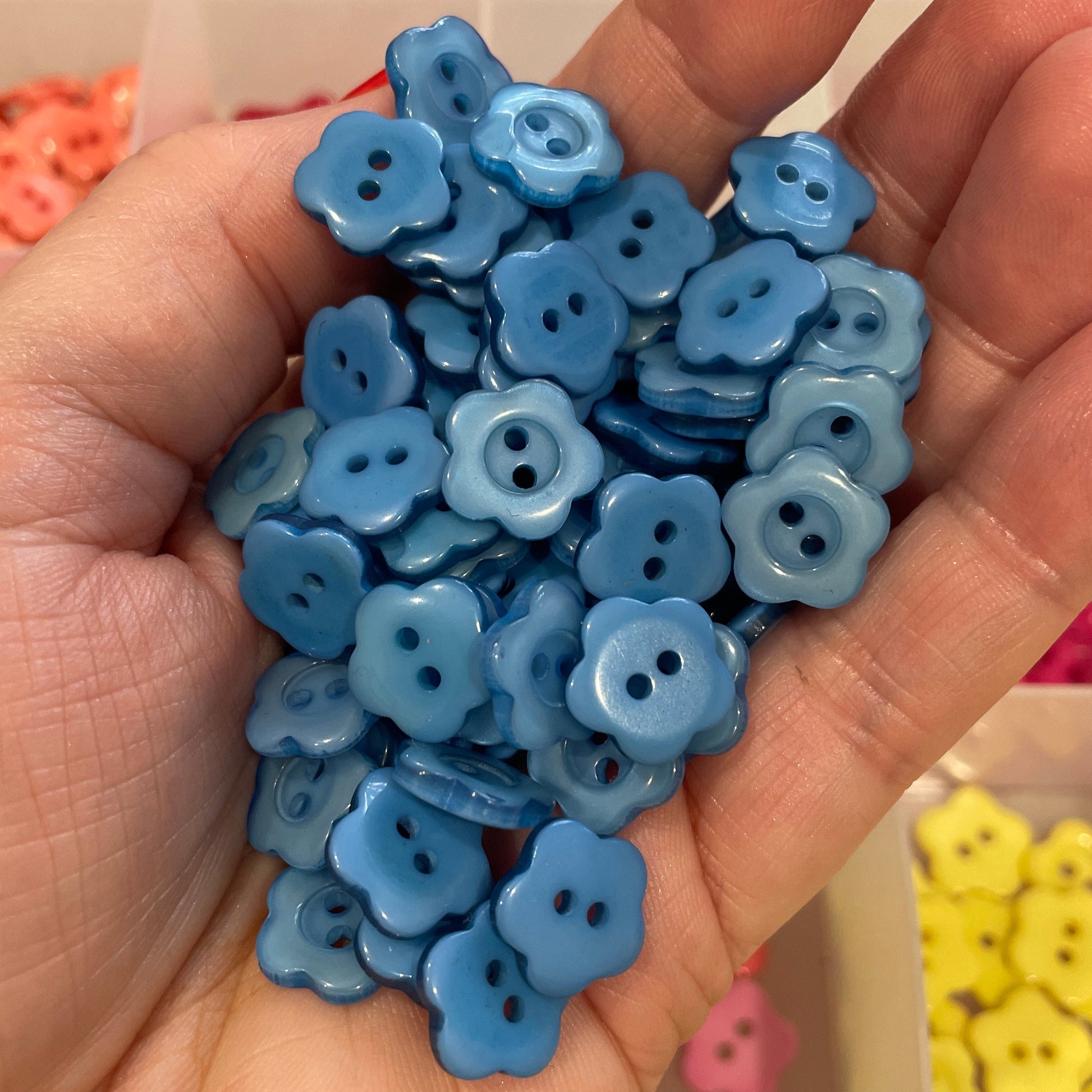 Hand holding blue flower-shaped buttons with a container of more buttons in the background.