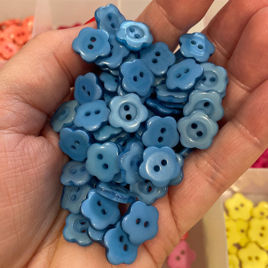 Hand holding blue flower-shaped buttons with a container of more buttons in the background.