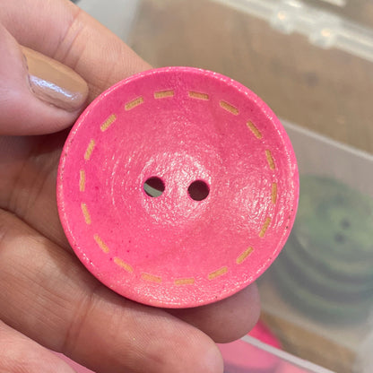 Pink button held by a hand with a container of pink buttons in the background.