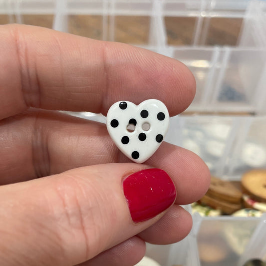 Hand holding a heart-shaped button with a container of various buttons in the background